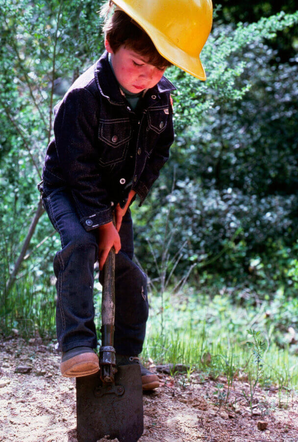 A young child wearing a yellow hard hat and denim jacket presses down on a shovel, digging in the dirt outdoors with greenery in the background.
