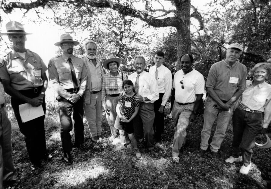 A group of people, including park rangers, adults, and a child, stand together outdoors under trees, smiling for the camera in black and white.