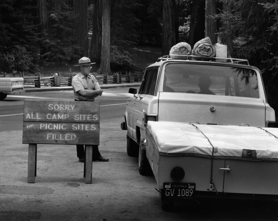 A park ranger stands by a sign reading “Sorry all camp sites and picnic sites filled” near a white SUV with camping gear on the roof and a trailer attached, surrounded by tall trees.
